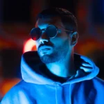 Artist SPE3D standing confidently in front of vibrant neon lights, wearing sunglasses.