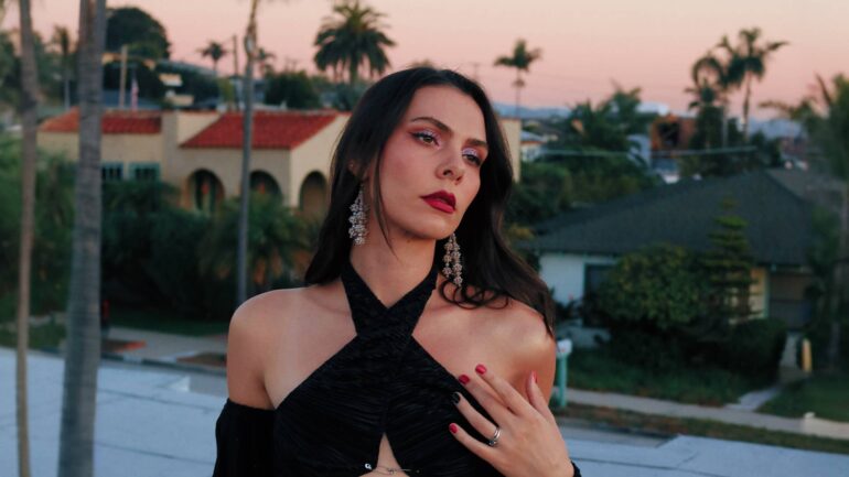 Estella Dawn in a stylish black dress with red lipstick, posing outdoors at dusk with palm trees in the background.