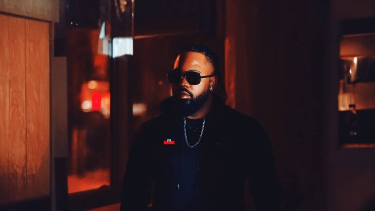 MARKUS BANKS in black hoodie with silver chain, dimly lit background with light reflections.