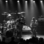 Band performs on stage, spotlighted in a monochrome concert setting.