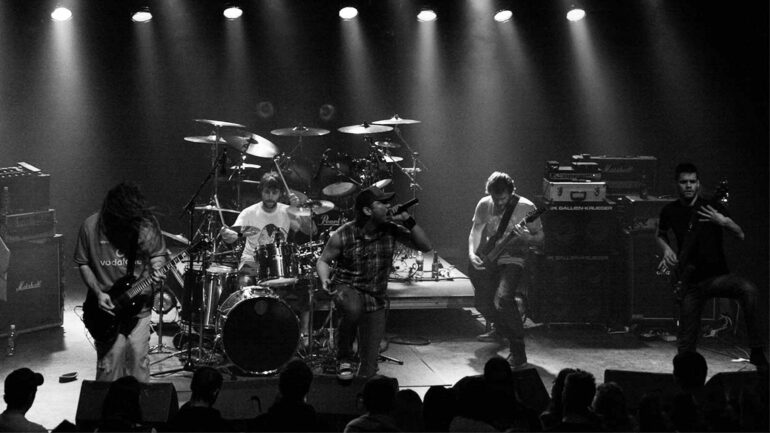 Band performs on stage, spotlighted in a monochrome concert setting.