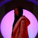 SAGEALINA in profile, wearing a textured jacket, against a glowing pink orb backdrop.