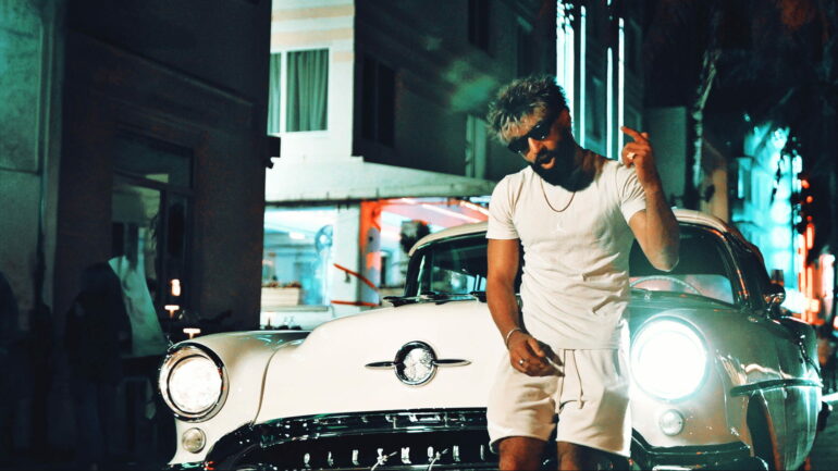 Artist Shubz stands by a classic car on a Miami street at night.