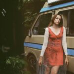 Singer-songwriter Sophie Kilburn stands beside a vintage van, clad in a red dress layered with a white lace top and a netted overlay.