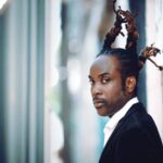 Reggae artist Nesbeth in a formal black jacket, standing by a wall.