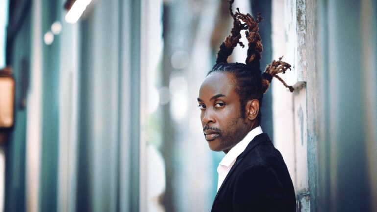 Reggae artist Nesbeth in a formal black jacket, standing by a wall.