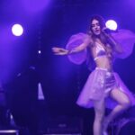 Juli Chan performs on stage during The Posh Tour, dressed in a lilac outfit, with bright vertical lights in the background.