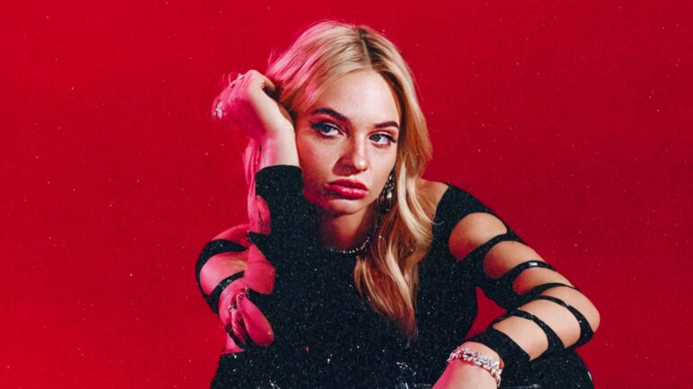 Cameron Sage From sitting on a red backdrop, posing with a black amplifier, wearing a black outfit with cut-out details.