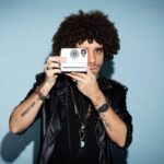 Jermaine Fleur holding a vintage camera, looking directly through the lens, with curly hair and a leather jacket.