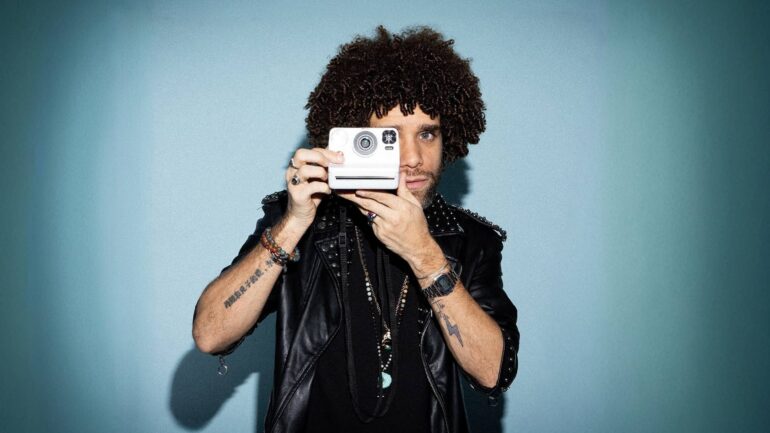 Jermaine Fleur holding a vintage camera, looking directly through the lens, with curly hair and a leather jacket.