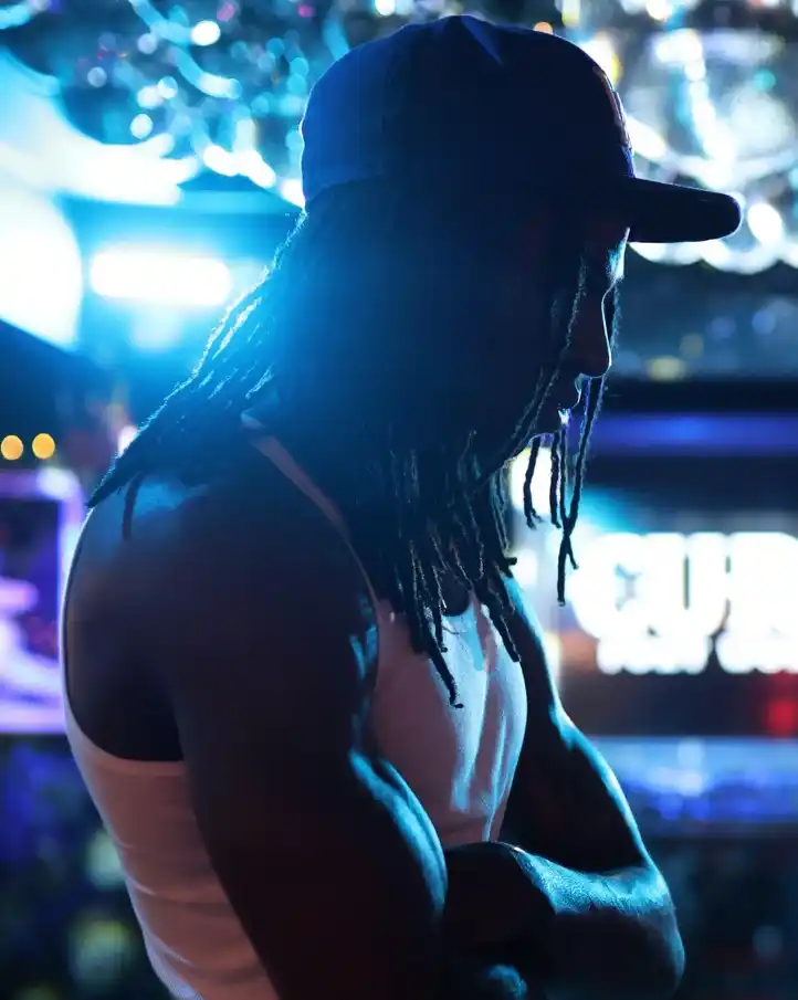 Mola stands with arms crossed, wearing a white tank top and cap, lit by blue and purple neon lights.