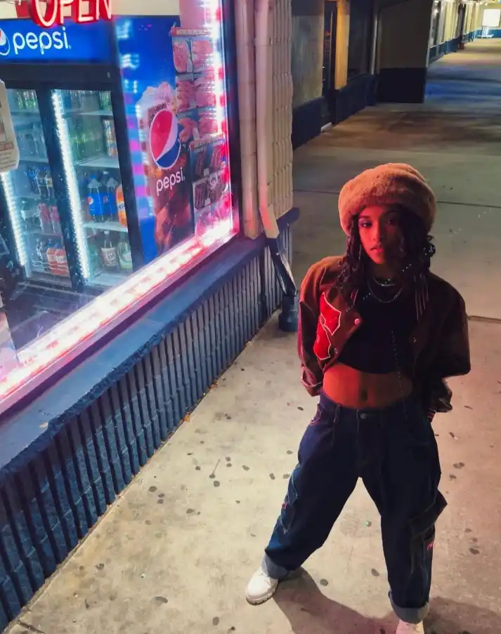 Tai Marie stands outside a store with bright neon signage, wearing a crop top, jacket, and baggy jeans.