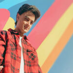 A-Zal posing in a red flannel shirt against a colorful striped background.