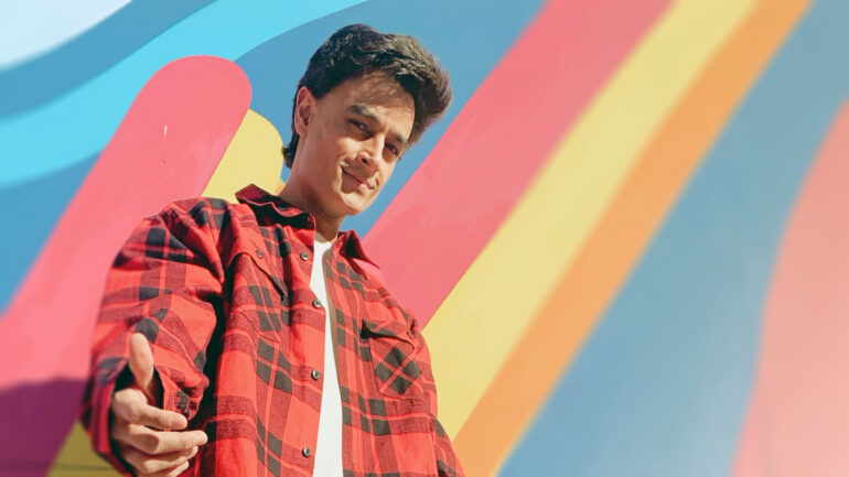 A-Zal posing in a red flannel shirt against a colorful striped background.