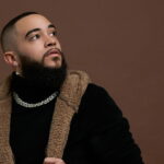 ALEXX sitting on a stool, looking up, dressed in a black outfit with a fur-lined jacket, against a brown background.