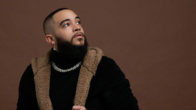 ALEXX sitting on a stool, looking up, dressed in a black outfit with a fur-lined jacket, against a brown background.