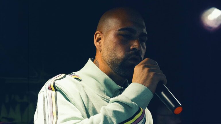 Dro Songz performing live, wearing a light-colored jacket with a microphone in hand.