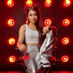 Gzelle C sits confidently in front of red lights, promoting her new single "Top Tier," which blends English and Spanish lyrics.