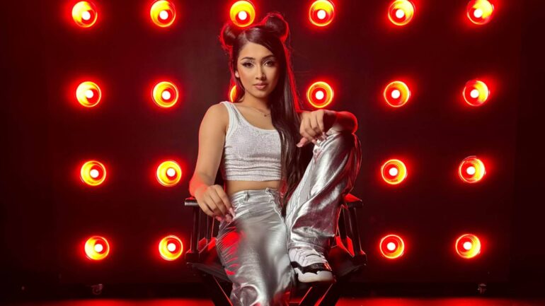 Gzelle C sits confidently in front of red lights, promoting her new single "Top Tier," which blends English and Spanish lyrics.