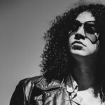 Black and white photo of Johnny Nasty Boots, a rock artist with curly hair and sunglasses, wearing a leather jacket, gazing into the distance.