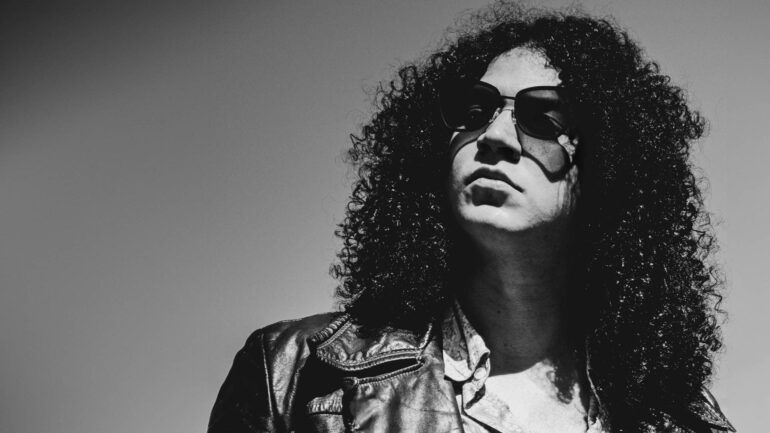 Black and white photo of Johnny Nasty Boots, a rock artist with curly hair and sunglasses, wearing a leather jacket, gazing into the distance.