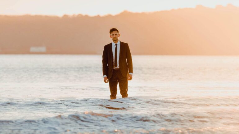 Josh Hearmon standing in the ocean in a suit at sunset, promoting his new single "Over Again."