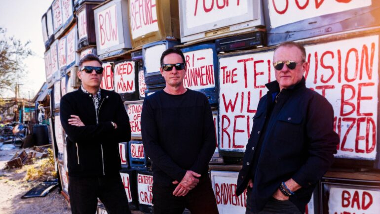 The band MAWK3 standing in front of a wall of televisions with provocative messages. Members Steve Giles, Jason Sweeny, and Dan Nelles are pictured.