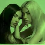 MYNX, the LA-based pop duo featuring Tori Wade and Valeria Arguelles, posing together with serious expressions against a green background.