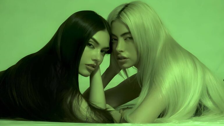 MYNX, the LA-based pop duo featuring Tori Wade and Valeria Arguelles, posing together with serious expressions against a green background.