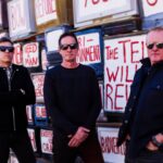 MAWK3 band members stand in front of a wall of old TVs with rebellious graffiti, promoting their new single "Masterpiece" released in August 2024.