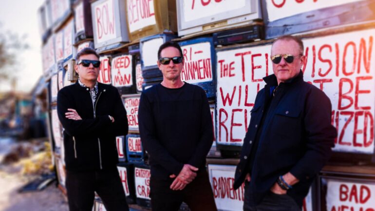 MAWK3 band members stand in front of a wall of old TVs with rebellious graffiti, promoting their new single "Masterpiece" released in August 2024.