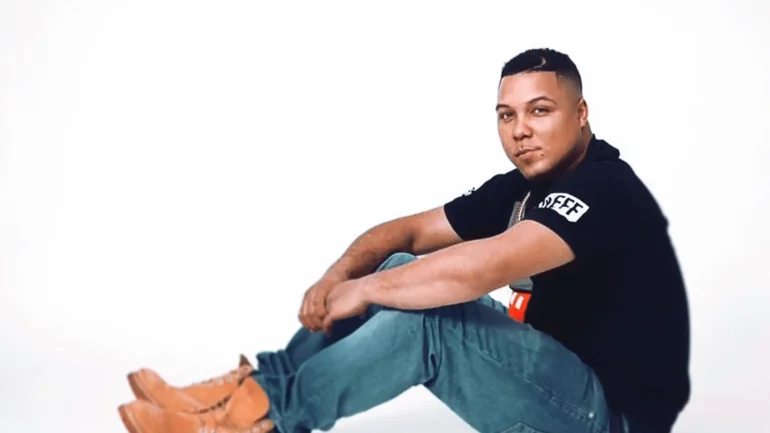 Diz Does sitting casually on the floor in a relaxed pose, wearing a black t-shirt, blue jeans, and tan boots, against a plain white background.