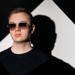 Oliver Sullivan, wearing sunglasses, stands in front of a dark background with a geometric shape, promoting his new hit 'Never Let You Go.'