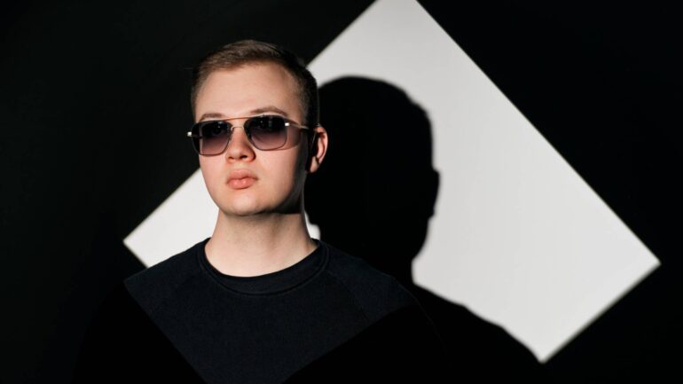 Oliver Sullivan, wearing sunglasses, stands in front of a dark background with a geometric shape, promoting his new hit 'Never Let You Go.'