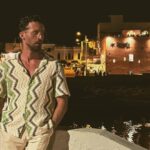 Rocco Bell' stands by a waterfront at night, dressed in a patterned shirt and beige trousers, reflecting his artistic and cultural influences. The photo was taken in a coastal town with dim lights in the background.