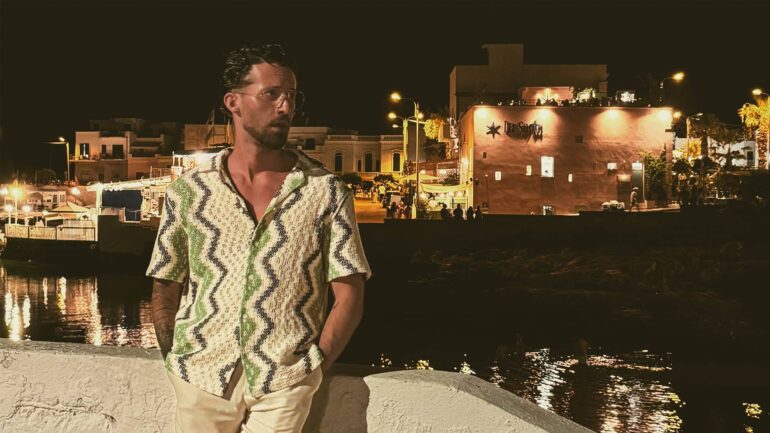 Rocco Bell' stands by a waterfront at night, dressed in a patterned shirt and beige trousers, reflecting his artistic and cultural influences. The photo was taken in a coastal town with dim lights in the background.