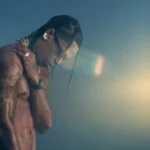 Travis Scott stands shirtless in a dimly lit scene, reflecting a moody and introspective moment from his latest music video “Drugs You Should Try It” celebrating 10 years of “Days Before Rodeo.”