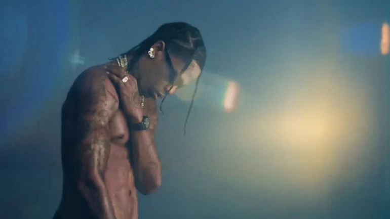 Travis Scott stands shirtless in a dimly lit scene, reflecting a moody and introspective moment from his latest music video “Drugs You Should Try It” celebrating 10 years of “Days Before Rodeo.”