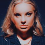 Bobbi Fallon posing in a dark, pinstripe blazer with a confident expression against a blue-lit backdrop.