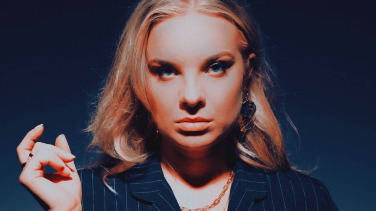 Bobbi Fallon posing in a dark, pinstripe blazer with a confident expression against a blue-lit backdrop.