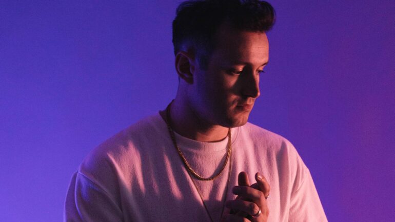 Drew Schueler posing against a purple background, hands clasped, capturing a moment of introspection.