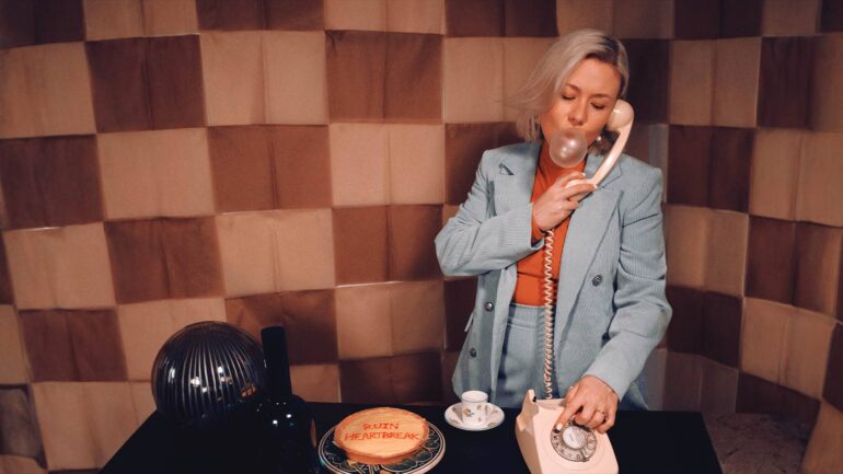 Hekla Goodman on the phone blowing a bubble, beside a cake labeled "Ruin Heartbreak."