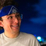 Man wearing a blue bandana and glasses, leaning on a railing with a city skyline in the background.