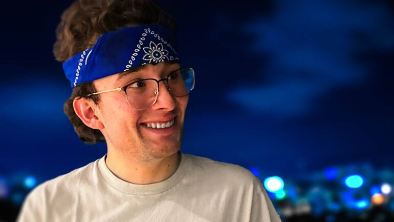 Man wearing a blue bandana and glasses, leaning on a railing with a city skyline in the background.