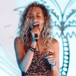 A woman with curly blonde hair sings into a microphone on stage, showcasing an energetic performance in a casual, sparkling top.