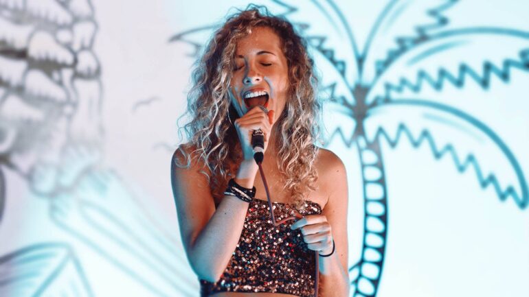 A woman with curly blonde hair sings into a microphone on stage, showcasing an energetic performance in a casual, sparkling top.