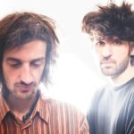 Two men with curly hair stand against a bright, overexposed background.