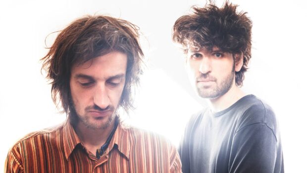 Two men with curly hair stand against a bright, overexposed background.