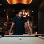 Dhan Aziz standing at a pool table, looking directly at the camera with a serious expression.