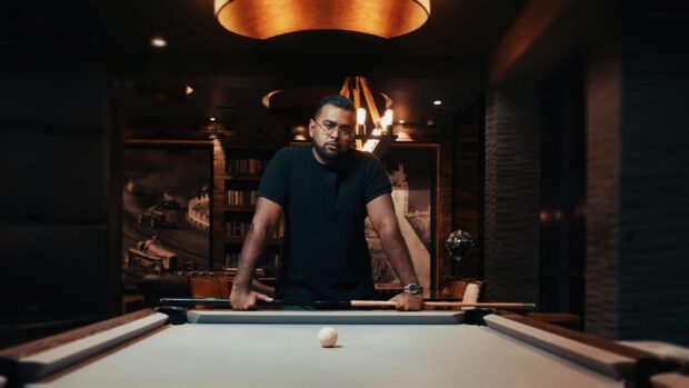 Dhan Aziz standing at a pool table, looking directly at the camera with a serious expression.